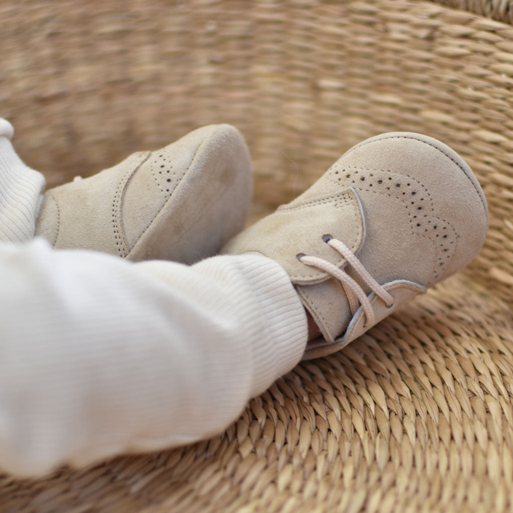 Close-up of a baby wearing Camel Suede Shoes from Baby Beau & Belle, resting on a woven basket. The baby is dressed in white knit pants. The handmade boys' shoes have decorative perforations on the sides and laces.