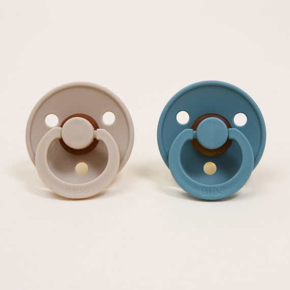 On a white background, two Bibs pacifiers are placed side by side. One is in vanilla and the other in island sea, both crafted from natural rubber. Each pacifier boasts a round shield with two ventilation holes and a circular handle embossed with "BIBS," offering a timeless and elegant touch that helps your little one self-soothe effortlessly.