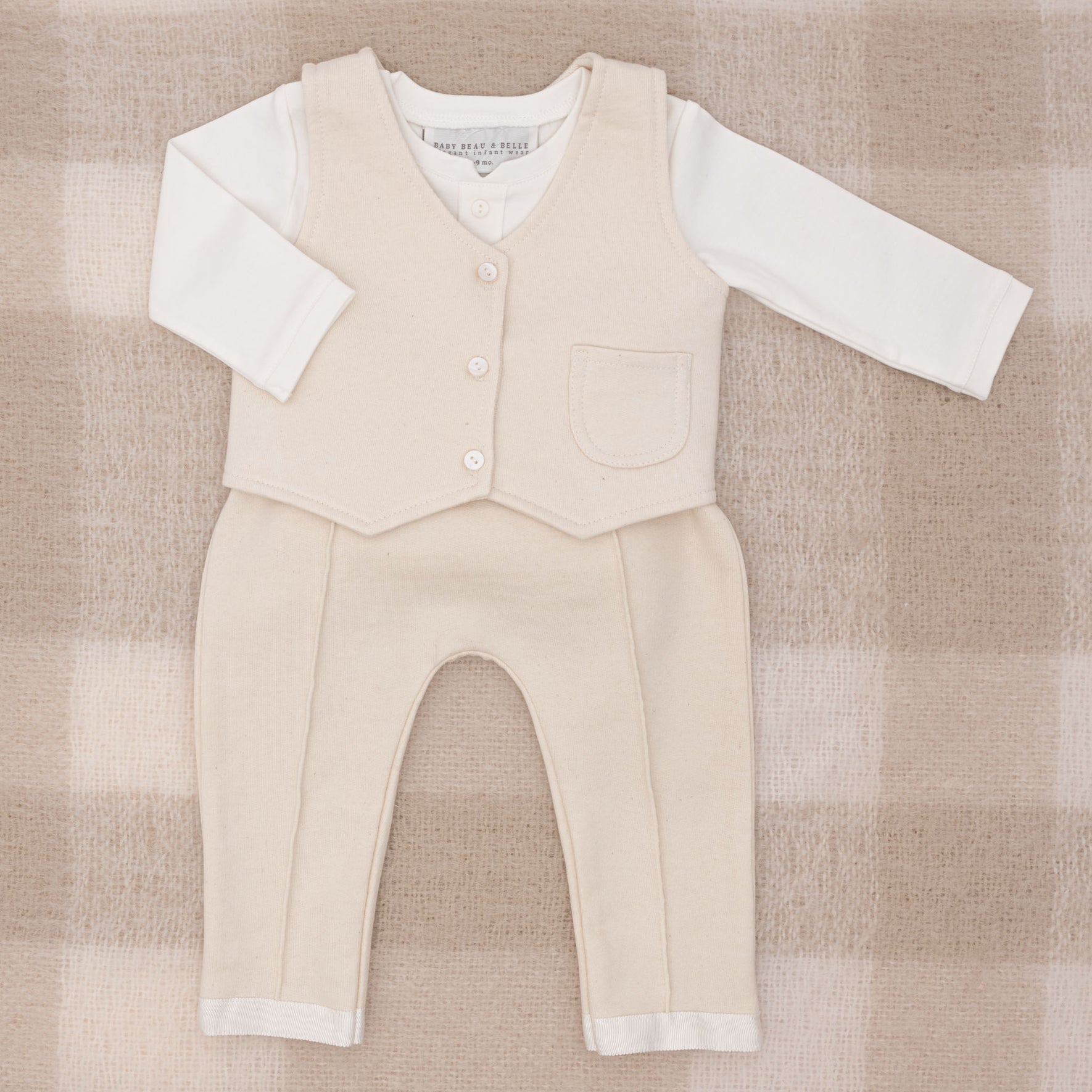 The Braden Vest Suit is a charming beige baby outfit perfect for any occasion. It features a long-sleeved white shirt paired with a beige vest adorned with buttons and a small pocket, along with matching pants. It is elegantly showcased on a soft, textured French Terry checkered background in beige and white.
