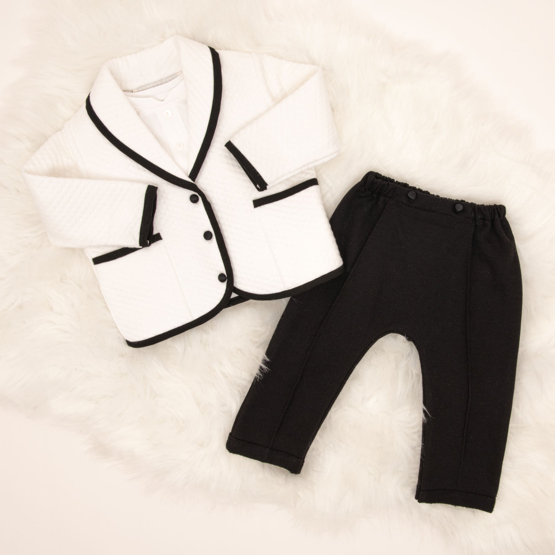 A flat lay of the James 3-Piece Suit, featuring the white quilted jacket with black trim and black pants, displayed on a soft fur background.