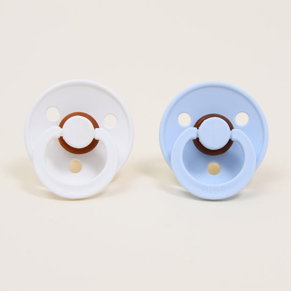 The Harrison Pacifier Set, featuring a white pacifier made from natural rubber for self-soothing and a light blue one, sits against a plain background. Both pacifiers have circular shields and handles.