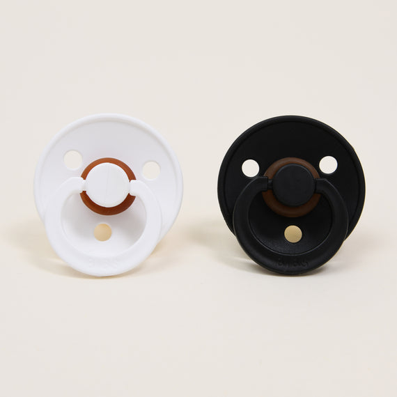 The Bibs Pacifier Set | White & Black features two natural rubber baby pacifiers displayed on a plain background. One pacifier is white with a brown center, and the other is black with a brown center. These Danish-designed pacifiers have circular shapes with handles to help babies self-soothe comfortably.