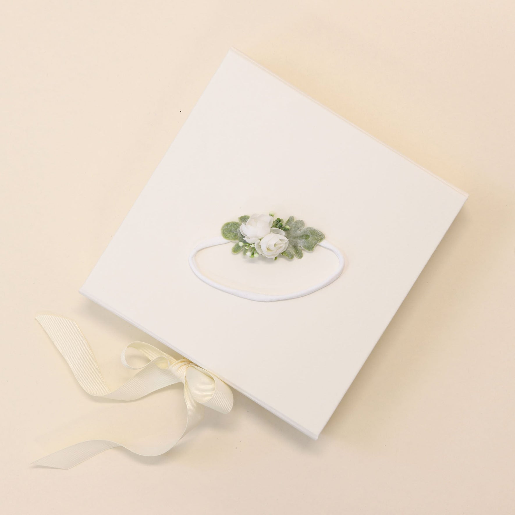 The Eliza Flower Headband is presented in a square white box adorned with a decorative floral design on the lid, showcasing two ivory flowers and green leaves. A cream ribbon elegantly ties the side of the box, which rests gracefully on a light beige surface, evoking the delicate craftsmanship of the handmade headband inside.