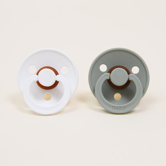 Two timeless and elegant baby pacifiers from the Bibs Pacifier Set in White & Sage are displayed against a plain backdrop. One pacifier is white, while the other is sage. Both feature circular shields with three ventilation holes and handle rings, crafted from natural rubber. The brand name "BIBS" is embossed on the sage pacifier.