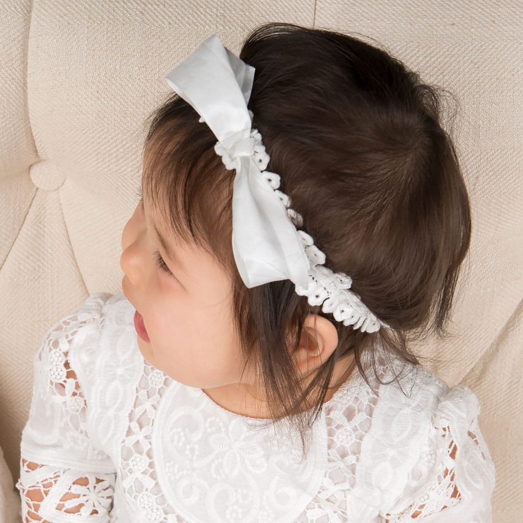 A toddler wearing an Adeline Accessory Bundle featuring a christening gown and a white bow headband looks to the side while seated on a cream-colored tufted chair.
