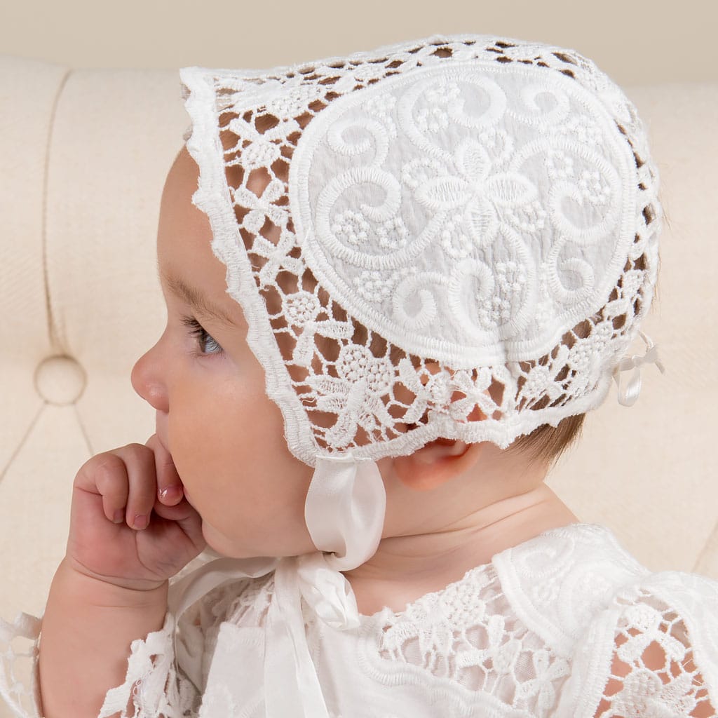 A baby wearing an Adeline Lace Christening Bonnet and matching baptism dress is shown in profile, gently sucking her thumb.
