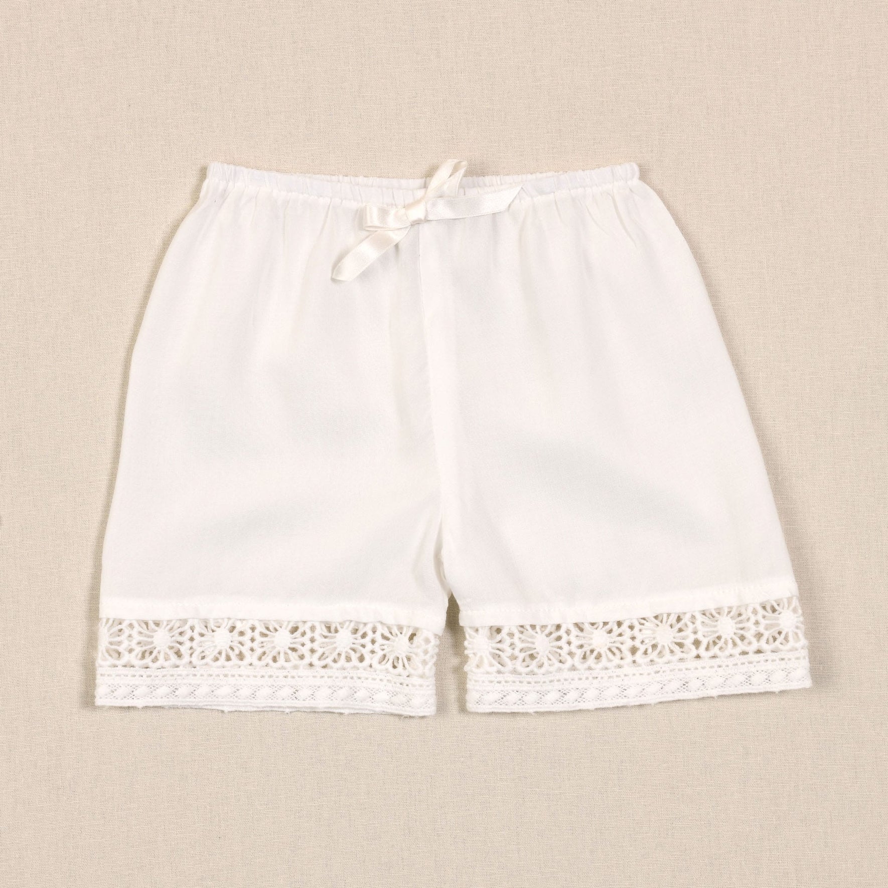 The Adeline Lace Christening Dress & Bloomers showcase a pair of white women's bloomers featuring an elastic waistband and adorned with a white silk ribbon sash tied in a bow at the front. These bloomers are elegantly detailed with intricate lace trim at the hem of each leg, echoing the classic design of a Christening Dress. The garment is displayed laid flat against a beige background.