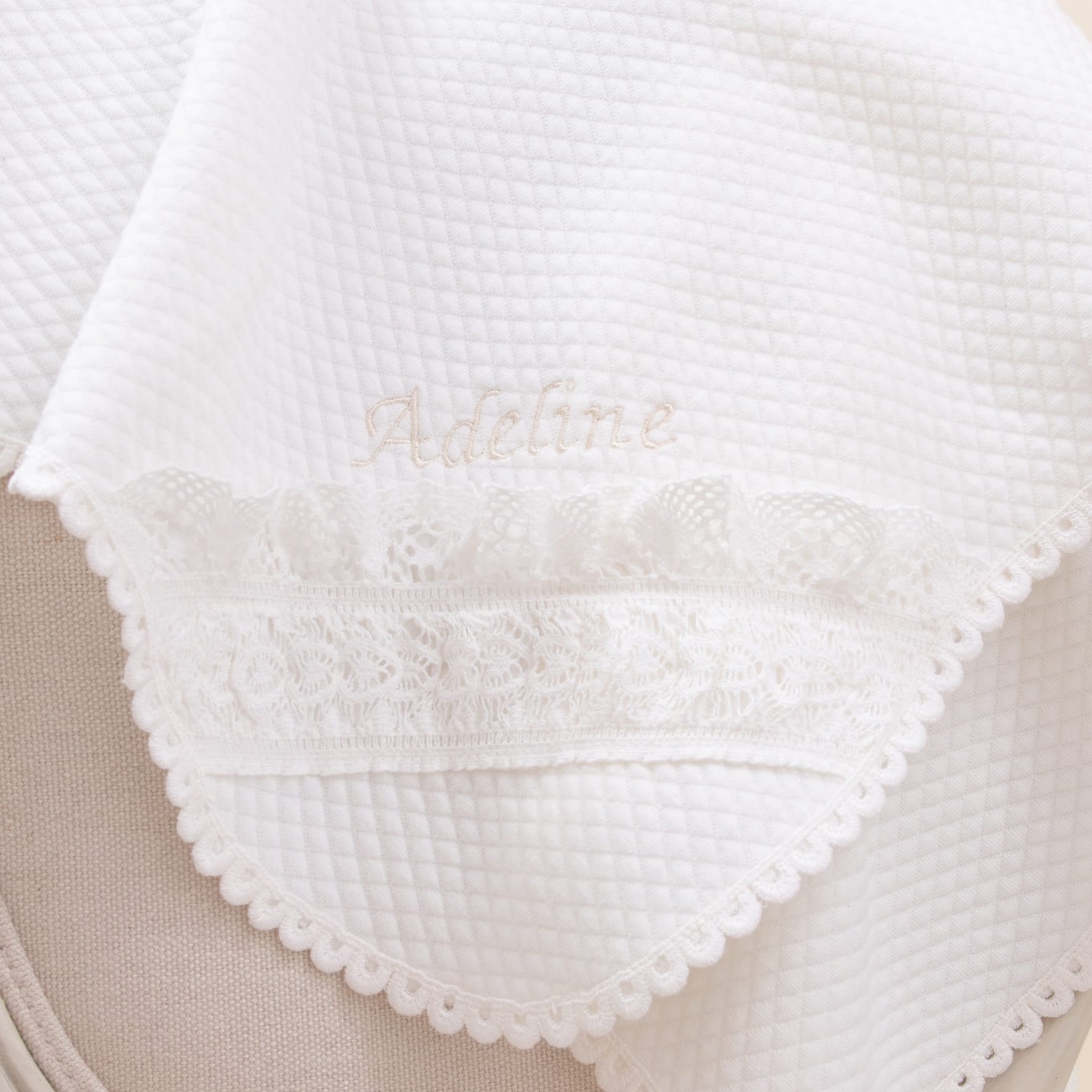 A close-up of the Adeline Personalized Blanket draped over a light-colored surface showcases its quilted cotton fabric, embroidered name "Adeline" in light beige, and delicate lace trim along the edge, making it a cherished keepsake.
