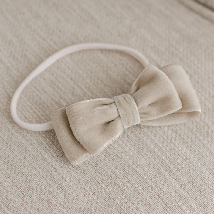 The Adeline Velvet Bow Headband, handmade in the USA, is displayed on a textured, light-colored fabric surface. Constructed from luxurious velvet ribbon, it showcases a neatly tied bow with a straightforward design that emphasizes its plush material and neutral beige hue.