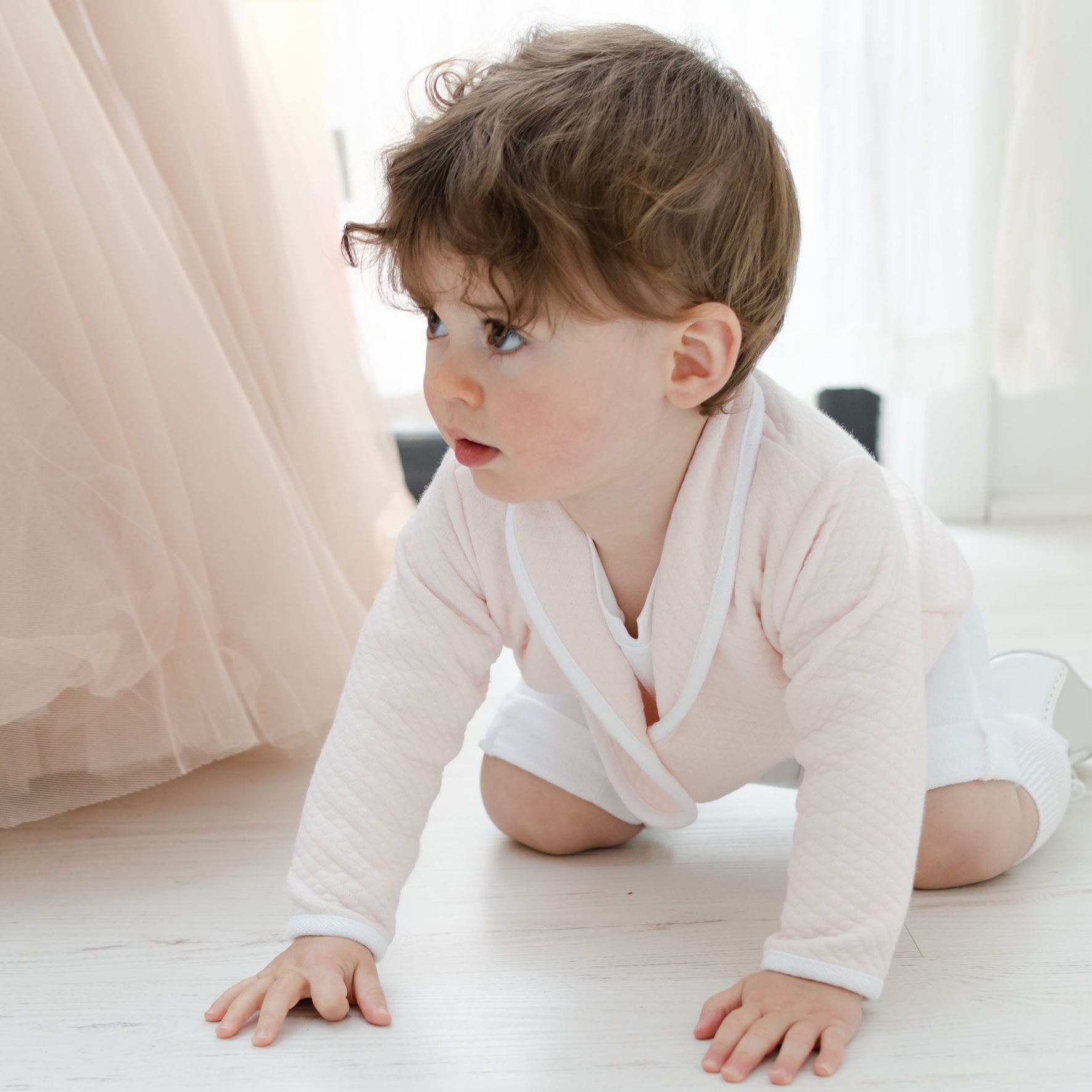 A toddler with brown hair crawls on a light-colored wooden floor, dressed in the Asher Blush 3-Piece Shorts Suit. The soft tulle fabric in the background hints at a special occasion outfit, perfectly completing this sweet moment.