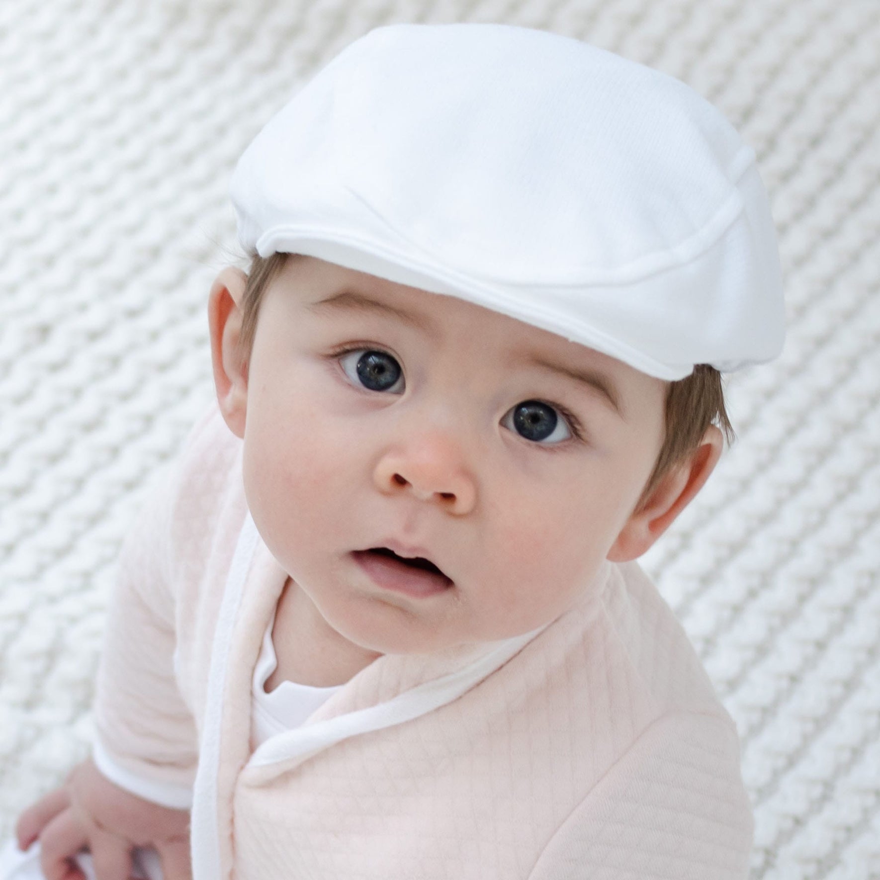 A baby wearing the Asher Blush 3-Piece Shorts Suit, complete with a white cap and blush shorts, gazes up at the camera. The backdrop consists of a textured, light-colored surface that enhances the scene's gentle charm.