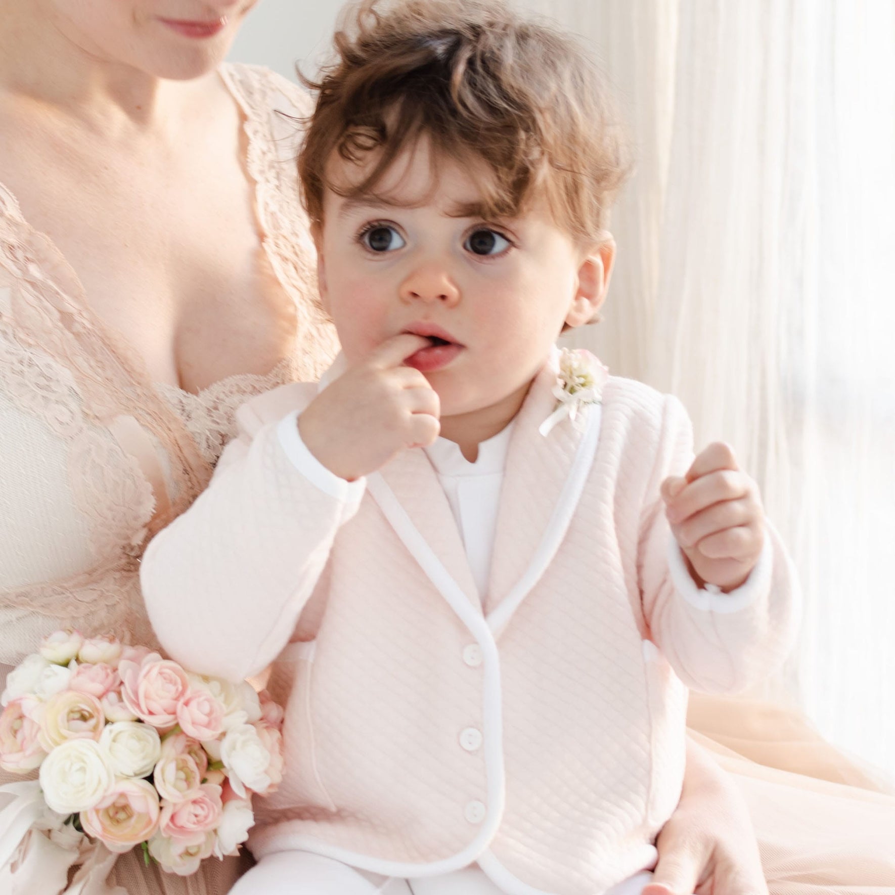 A woman is seated indoors with a child wearing the Asher Blush 3-Piece Shorts Suit, featuring a pale pink jacket and blush baby shorts. The curly brown-haired child looks to the side with a finger in their mouth. For this special occasion, the woman holds a small bouquet of light pink and white flowers.