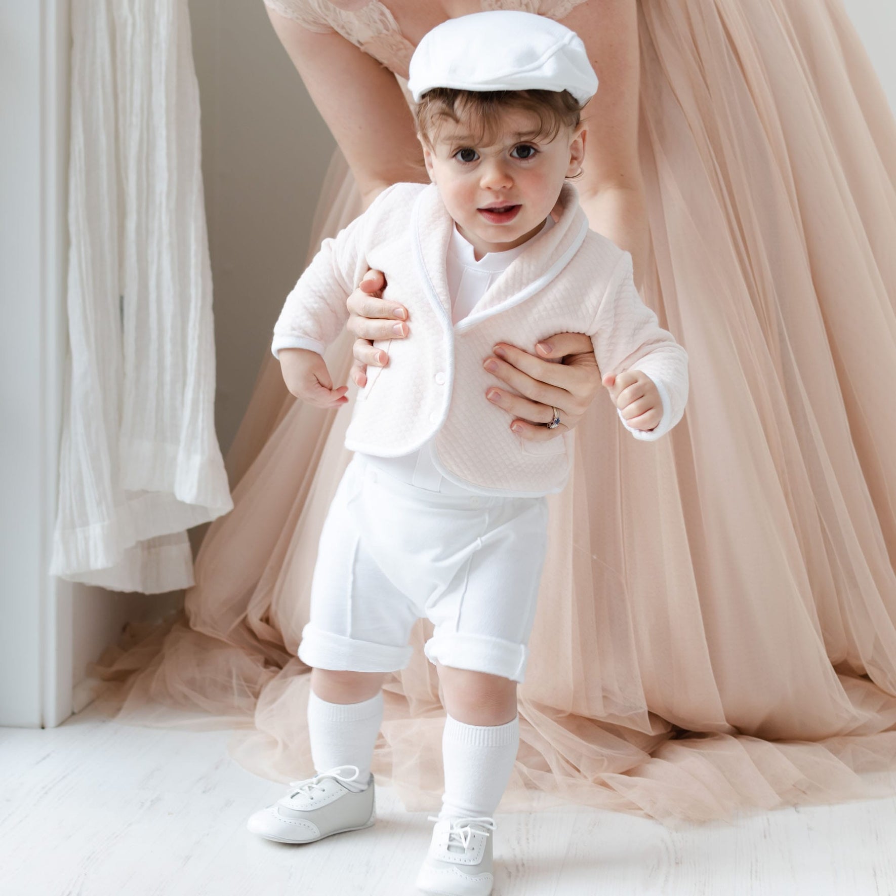 A young child dressed in the Asher Blush 3-Piece Shorts Suit, complete with a white hat and quilted cotton jacket, stands indoors. An adult, partially visible, supports the child from behind in a brightly lit environment with a soft, light-colored floor.