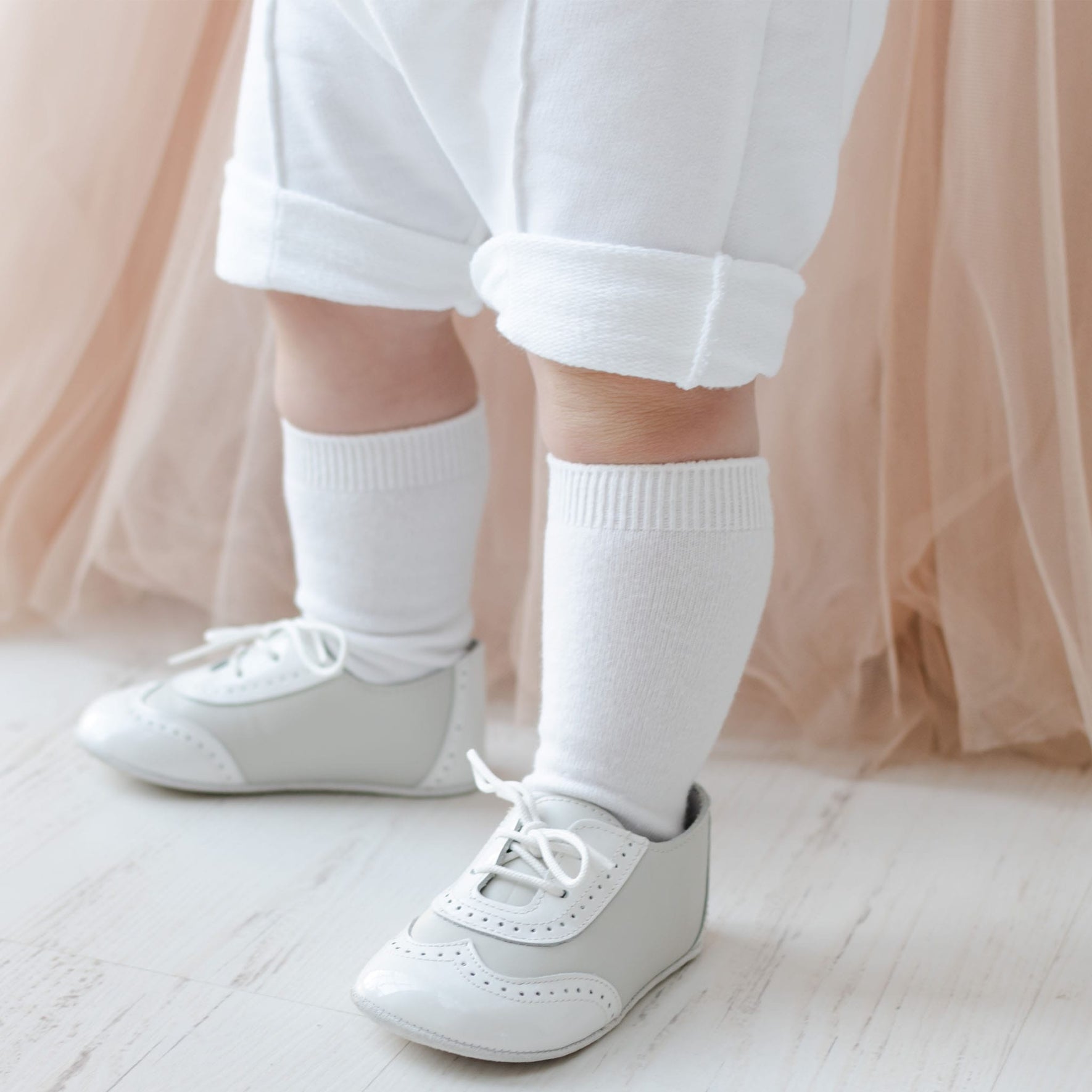The image focuses on a child's legs and feet dressed in the Asher Blush 3-Piece Shorts Suit, which includes white rolled-up pants, long white socks, and light gray lace-up shoes. The child stands on a light-colored wooden floor with a beige, tulle-like fabric softly cascading in the background.