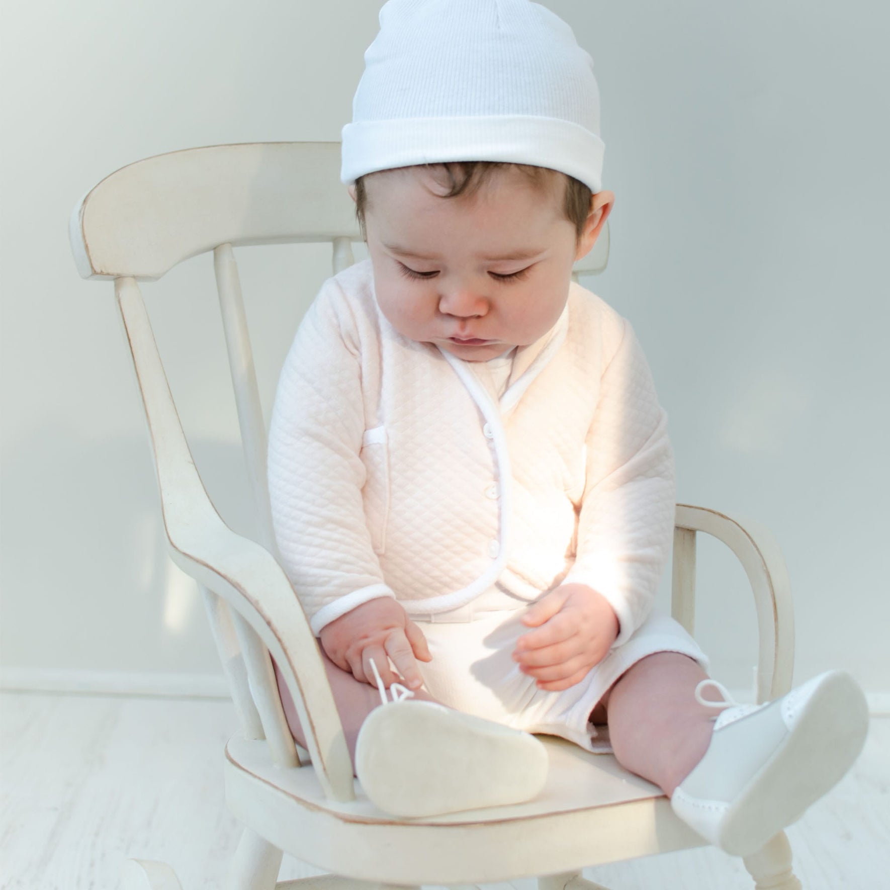 Dressed in an Asher Blush 3-Piece Shorts Suit, a baby sits on a white wooden chair, complete with a white hat and blush shorts. The softly lit scene captures the baby's gentle gaze down at its shoes, creating a peaceful atmosphere.