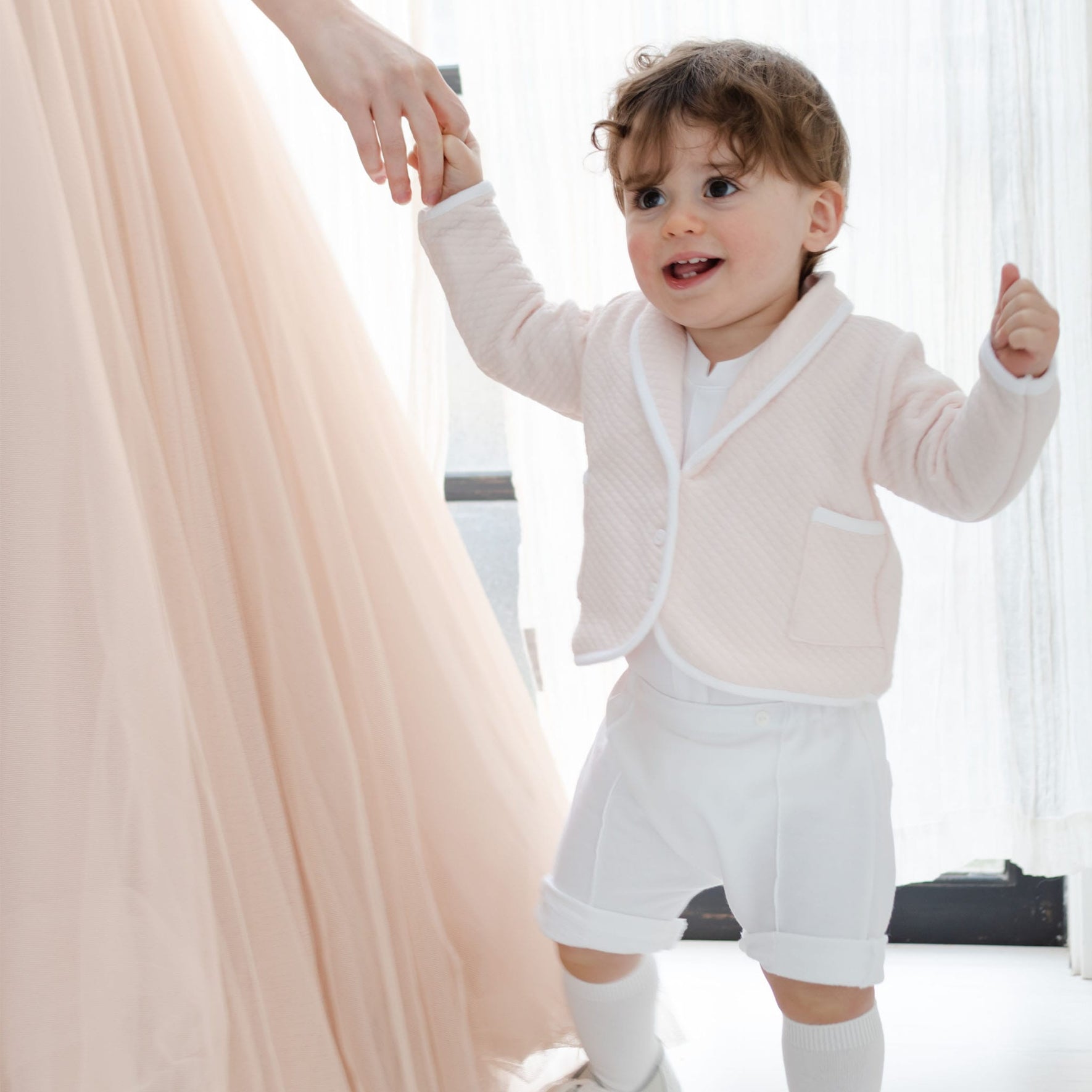 A young child in a special occasion attire, featuring the Asher Blush 3-Piece Shorts Suit with a quilted cotton jacket and blush baby shorts, holds an adult's hand. The adult is clad in a long, flowing dress. They appear to be indoors, standing by a light-filled window.