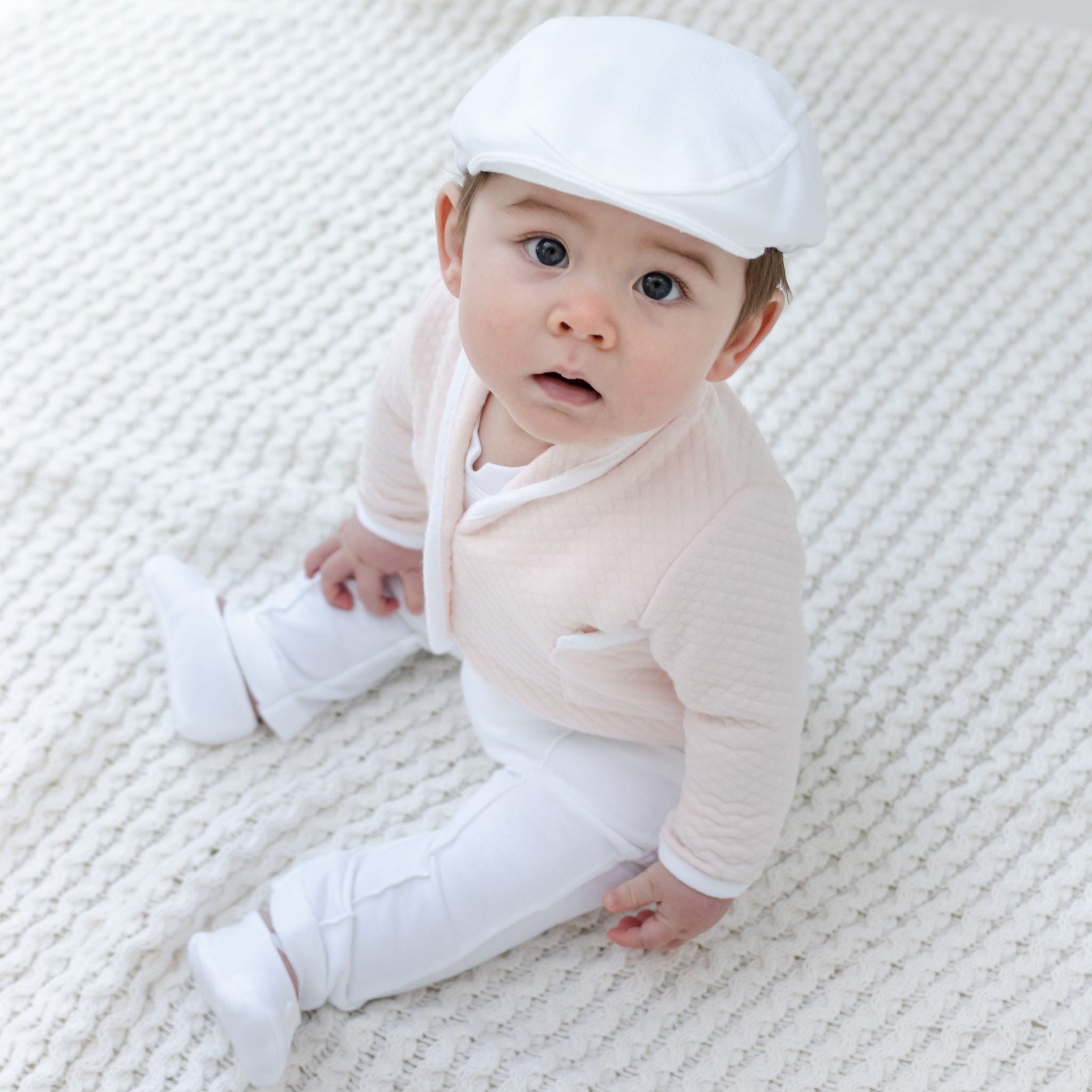 A baby, dressed in an Asher Blush 3-Piece Suit consisting of a baby suit with a white cap and light pink jacket, sits on a textured cream-colored blanket. The little one is also wearing cozy white pants and socks, looking up with an inquisitive expression.