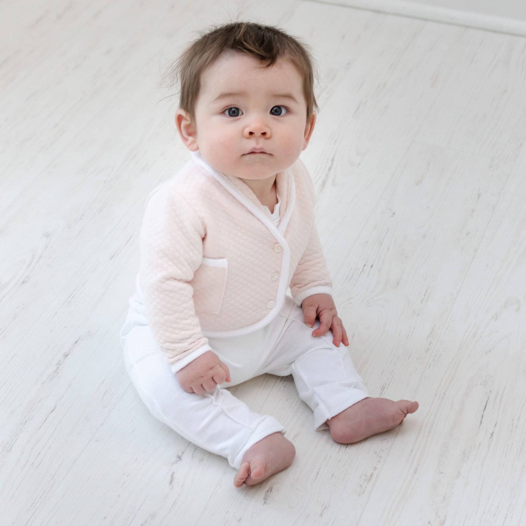 A baby with light brown hair and blue eyes sits on a light wooden floor, dressed in an Asher Blush 3-Piece Suit that includes a soft pink cardigan. The quilted cotton of the newborn outfit adds texture to the peaceful setting against the simple white background.
