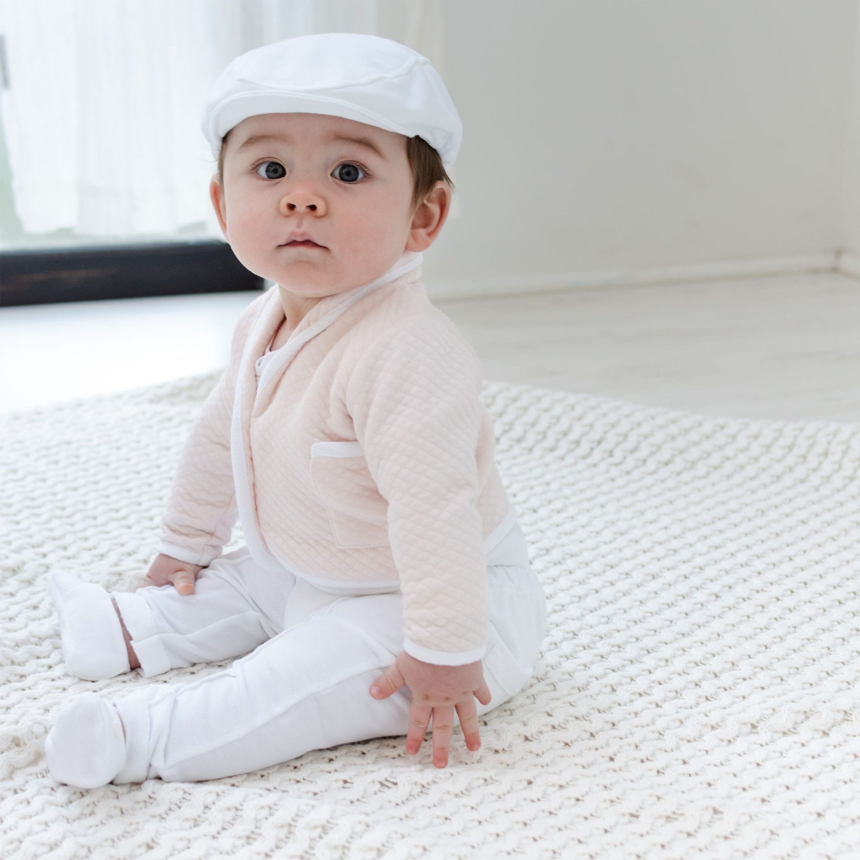 A baby wearing an Asher Blush 3-Piece Suit along with a white hat is seated on a knitted blanket on the floor. The room, characterized by its soft lighting and minimal decor, exudes a serene ambiance.