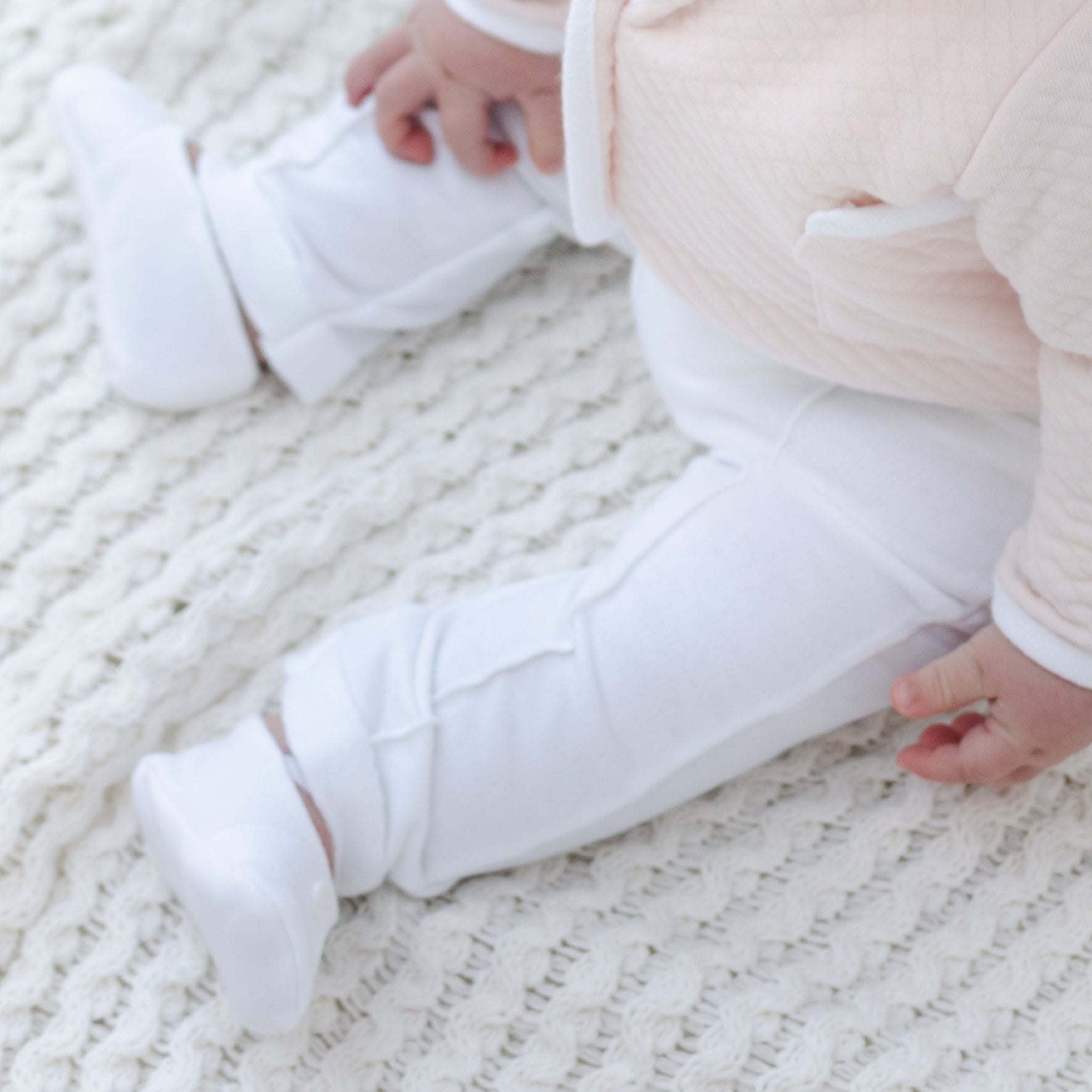 A baby wearing the Asher Blush 3-Piece Suit, which includes a pale pink jacket and white pants, is resting on a textured cream blanket. The image showcases the newborn's legs and feet adorned in matching white shoes.