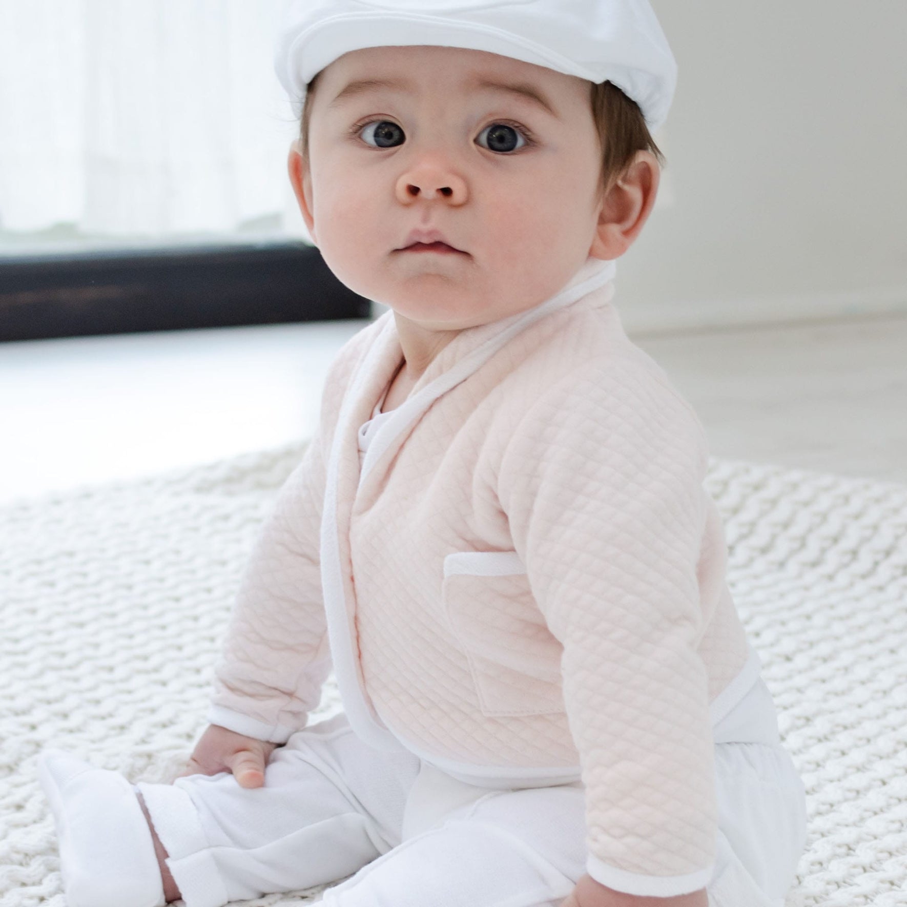 A baby wearing an Asher Blush 3-Piece Suit and cap is seated on a quilted cotton blanket. With a curious expression, the little one is comfortably positioned indoors, with a soft-focus window providing the background.