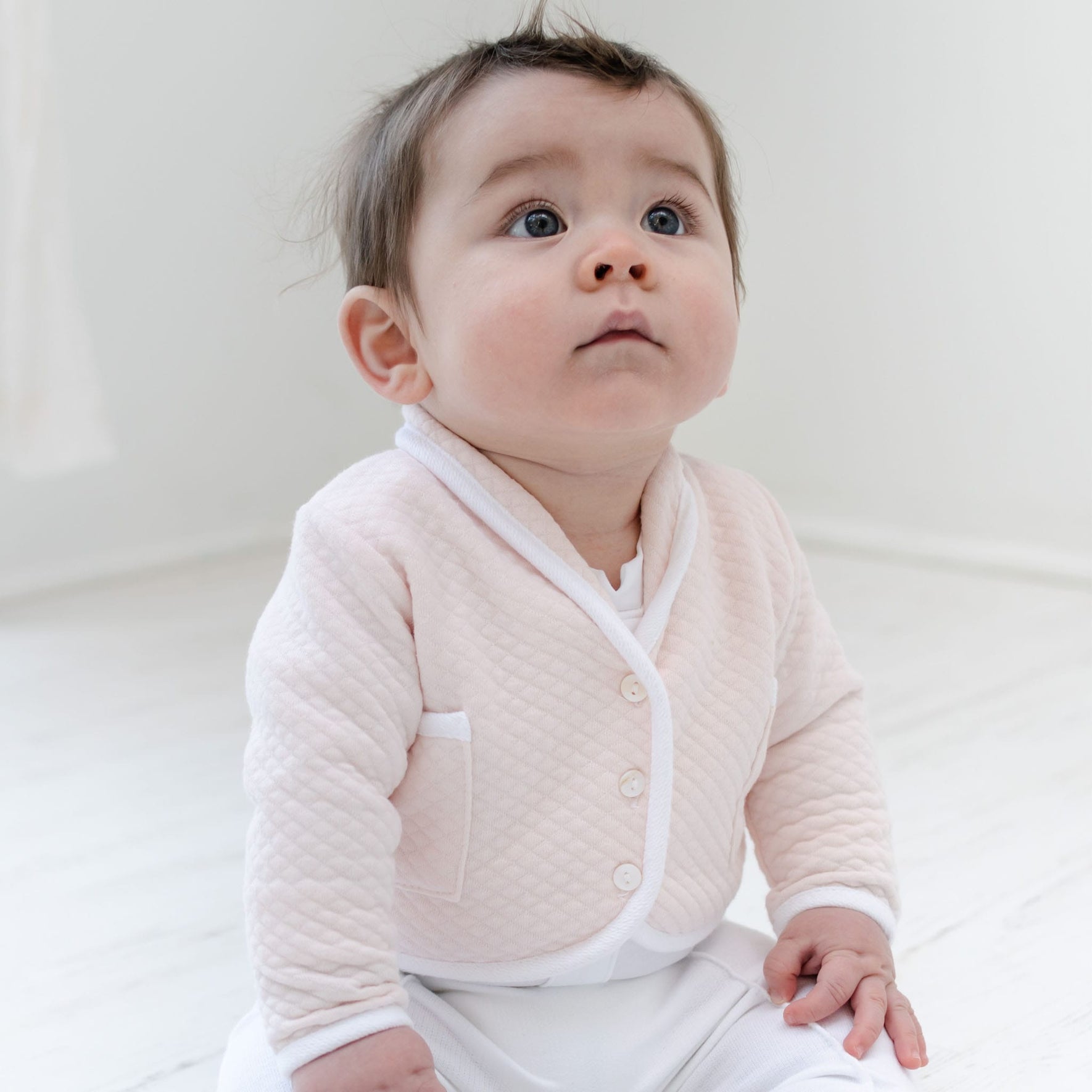 Wearing an Asher Blush 3-Piece Suit, which includes a quilted pink jacket with white trim, a baby sits on a light-colored floor, gazing upward. With short brown hair and wide eyes, the infant is framed by a softly lit and minimalistic background, capturing a serene newborn moment.