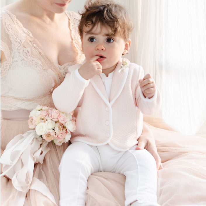 A woman in a light pink lace dress holds a toddler wearing the Asher Blush 3-Piece Suit, featuring a pale pink jacket and white pants, with the little one sucking on their fingers. She holds a bouquet of pink and white roses as they sit in a softly lit room with sheer curtains.
