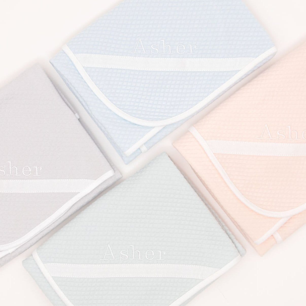 Flat lay photo of the four available colors of the Asher Personalized Blanket (folded in the photo), including grey, pink, powder blue, and soft teal. The blanket is made out of a 100% Quilted Cotton and features an embroidered name on the corner of the blanket.