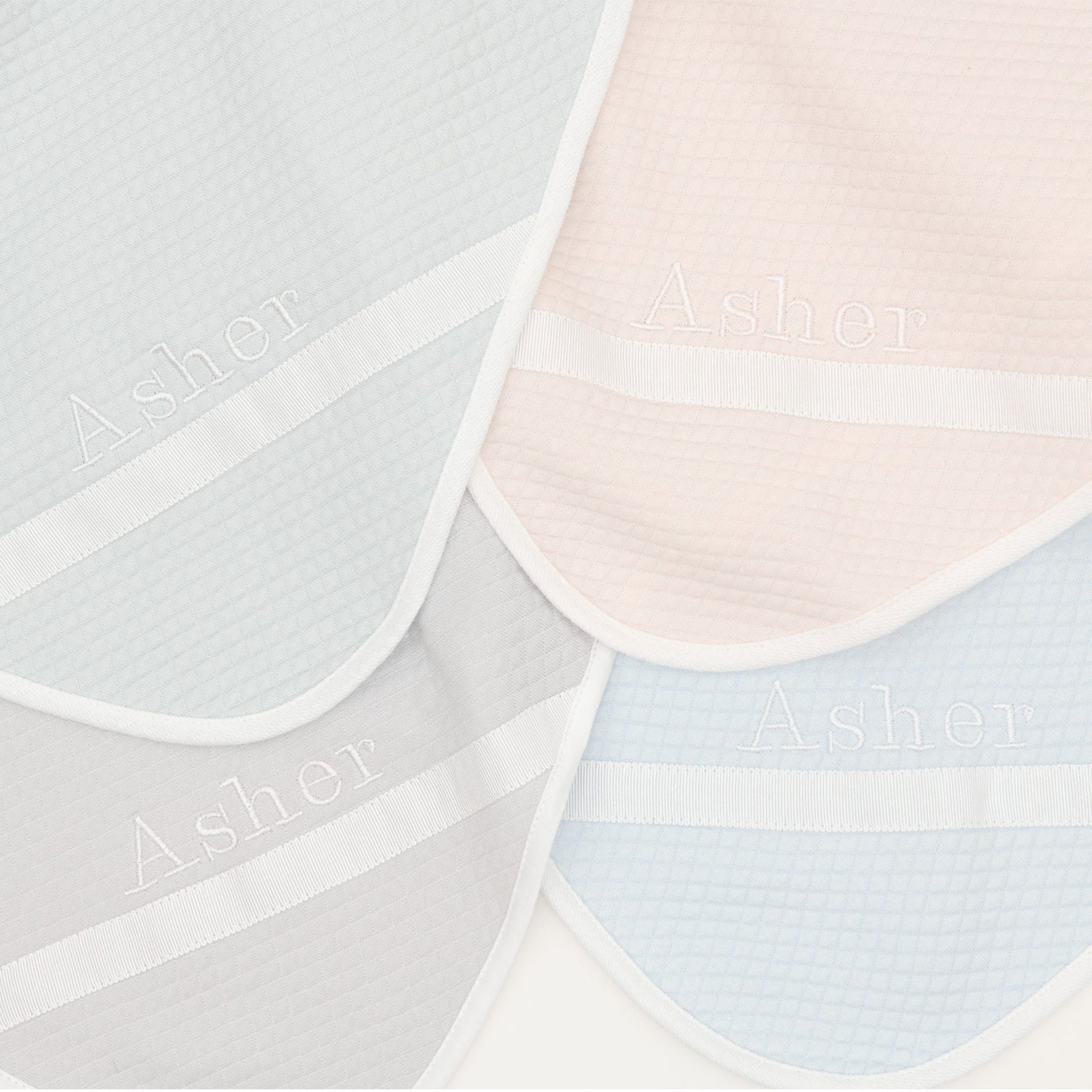 Flat lay photo of the four available colors of the Asher Personalized Blanket, including grey, pink, powder blue, and soft teal. The blanket is made out of a 100% Quilted Cotton and features an embroidered name on the corner of the blanket.