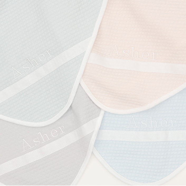 Flat lay photo of the four available colors of the Asher Personalized Blanket, including grey, pink, powder blue, and soft teal. The blanket is made out of a 100% Quilted Cotton and features an embroidered name on the corner of the blanket.