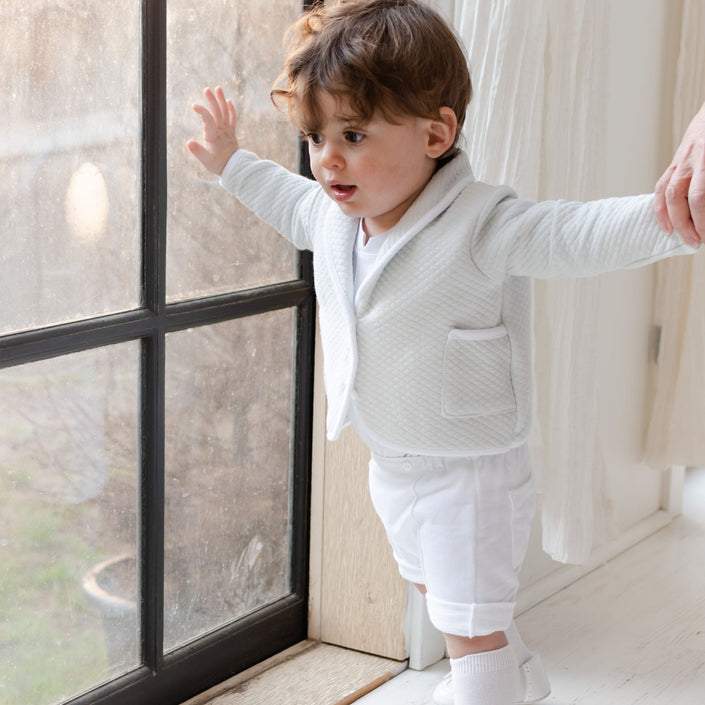 A young child stands by a window, dressed in a special occasion outfit featuring the Asher Soft Teal 3-Piece Shorts Suit. The child's hands are held by an unseen individual, and the room's white walls and curtains add to the tender scene.