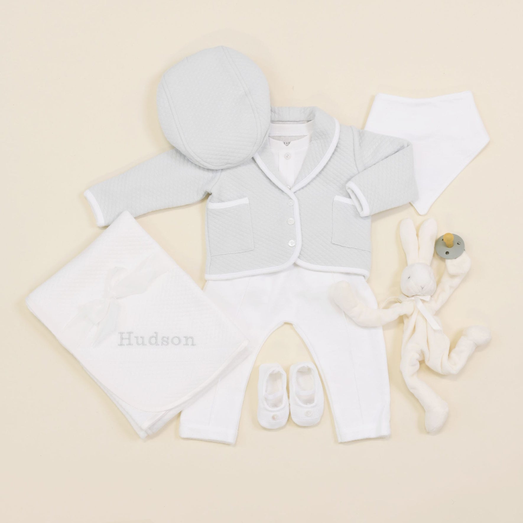 The Asher Soft Teal 3-Piece Suit includes a cozy light gray quilted cotton jacket, a matching hat, white pants, and small booties. Additionally, it comes with a soft white blanket featuring "Hudson" embroidery and a stuffed rabbit toy with a rattle.
