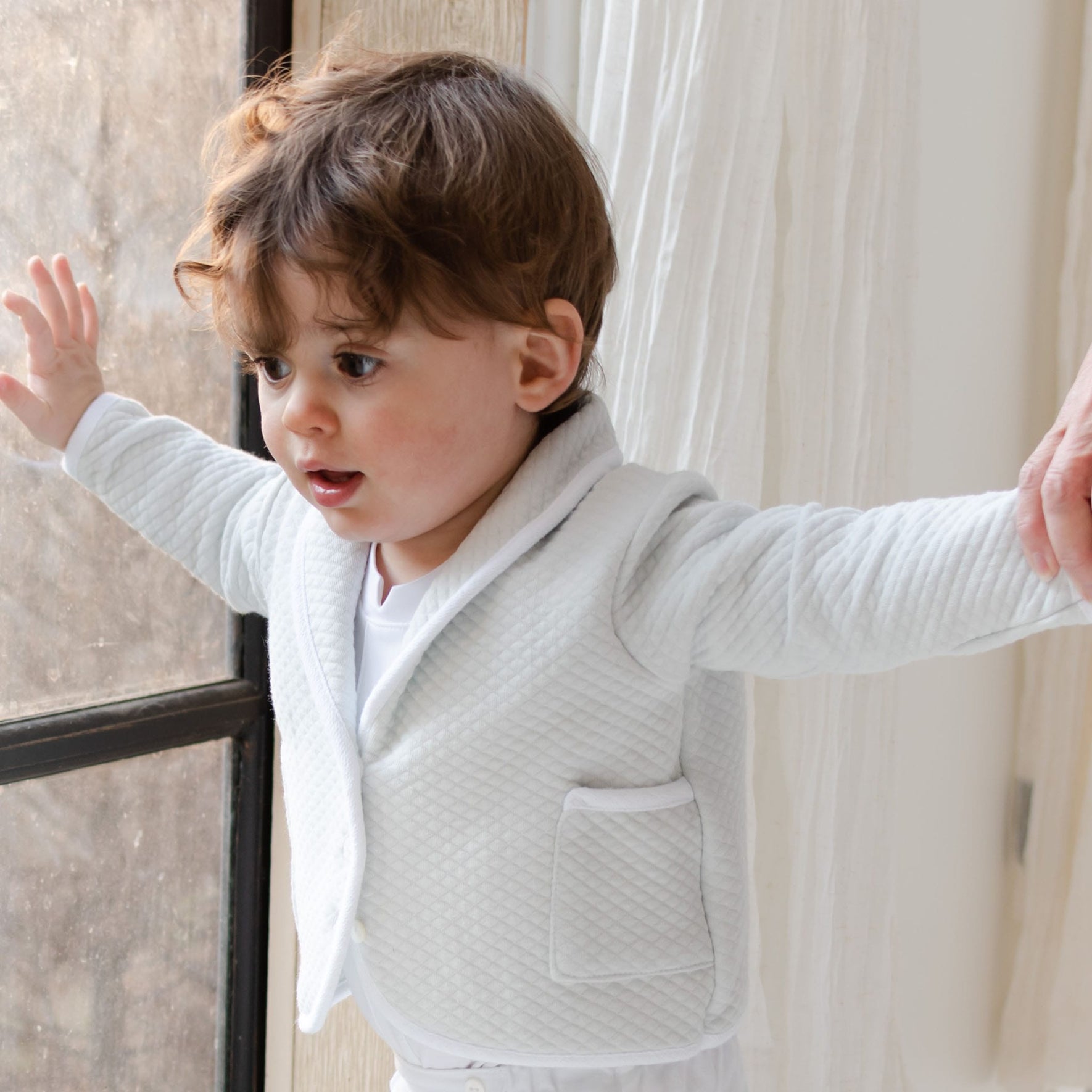 A toddler with curly hair, wearing the Asher Soft Teal 3-Piece Suit's quilted cotton jacket, holds onto a person's hand while leaning against a window. The setting appears to be indoors with natural light streaming in.