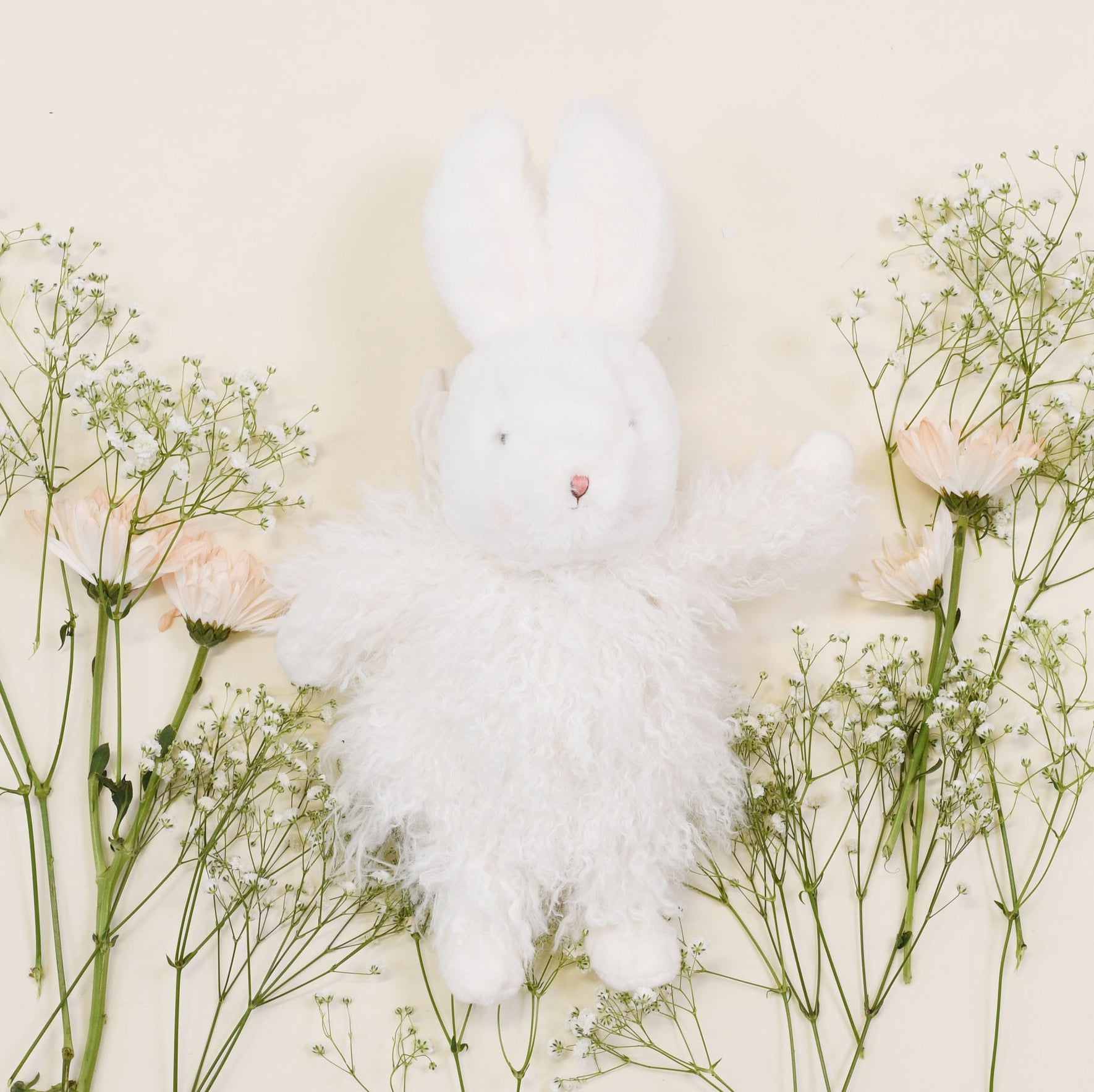 The Aurora Bunny Friend, featuring soft white shaggy fur, is beautifully surrounded by delicate baby's breath and pale pink flowers on a light cream background, making it an ideal baby shower gift.