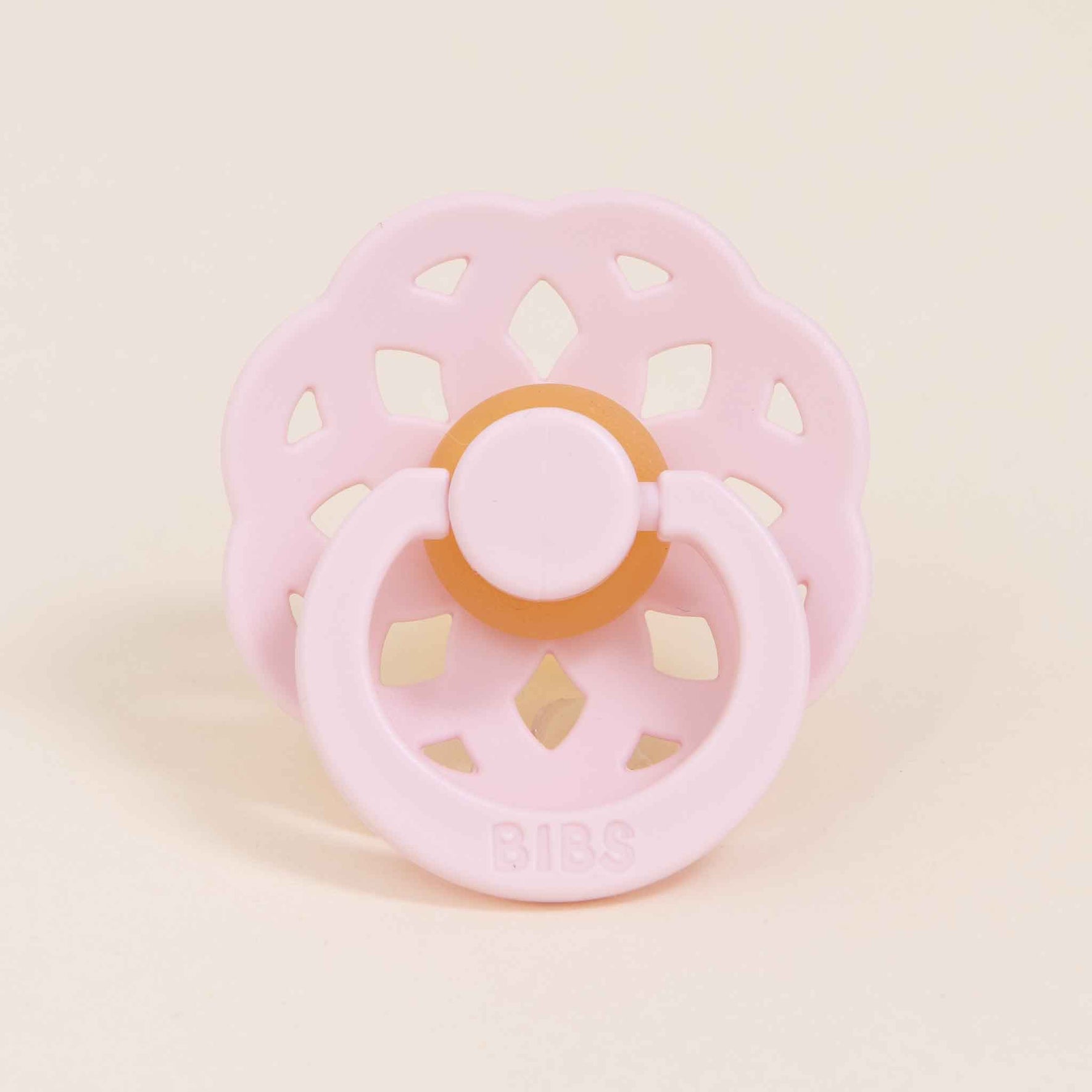 A pink pacifier from the Bibs Lace Pacifier 2 Pack in the Blossom color is featured against a plain background. This Danish-crafted pacifier, made from natural rubber, showcases a flower-shaped guard and an orange ring with the "BIBS" brand name embossed on the handle, designed to help your baby self-soothe effortlessly.