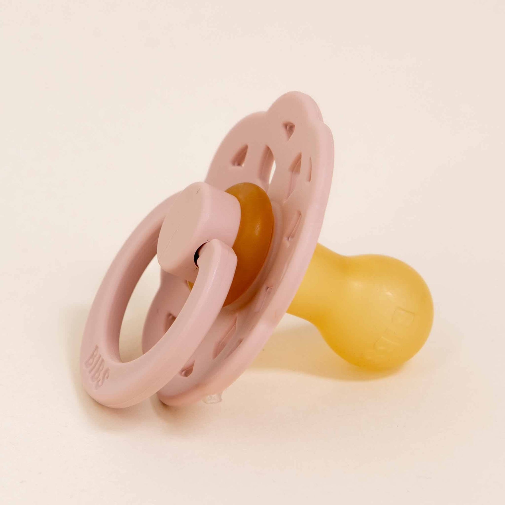 A Blush Bibs Lace Pacifier from a 2-pack set rests on a light-colored surface, showcasing its bohemian design with a perforated shield. The nipple is made of natural rubber, and the handle and shield display vibrant pink hues.