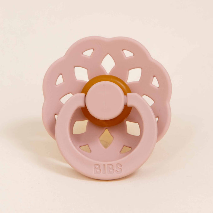 A Bibs Lace Pacifier in Blush, designed with a flower-shaped, bohemian style featuring circular cutouts, is showcased against a neutral background. Made from natural rubber, it has the word "BIBS" embossed on the handle to stylishly assist your baby in self-soothing.