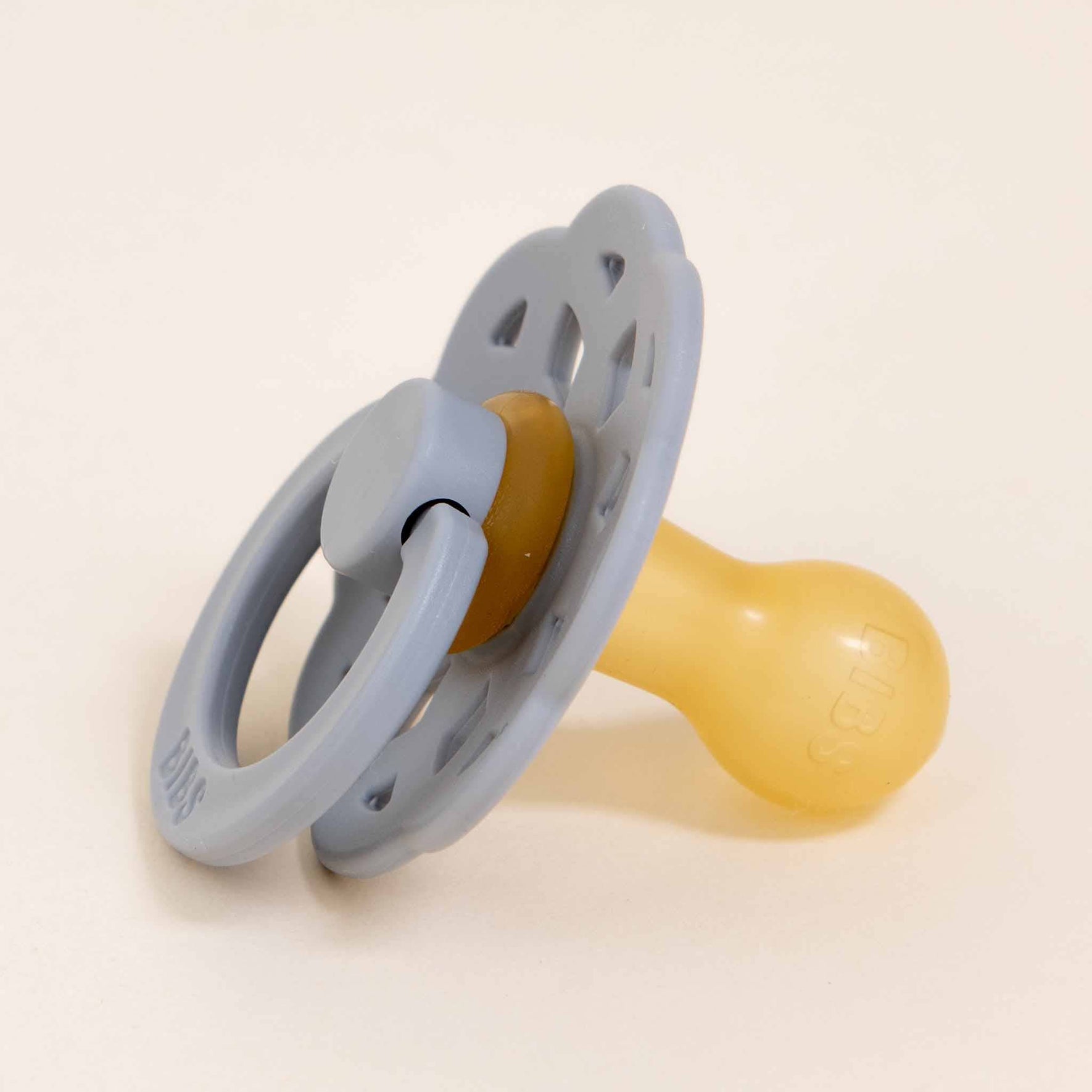 A Bibs Lace Pacifier from the 2-pack set named Cloud, featuring a blue perforated shield and handle loop with a yellow natural rubber nipple, is resting on a light surface. Designed to help infants self-soothe, the pacifier combines blue and yellow hues.