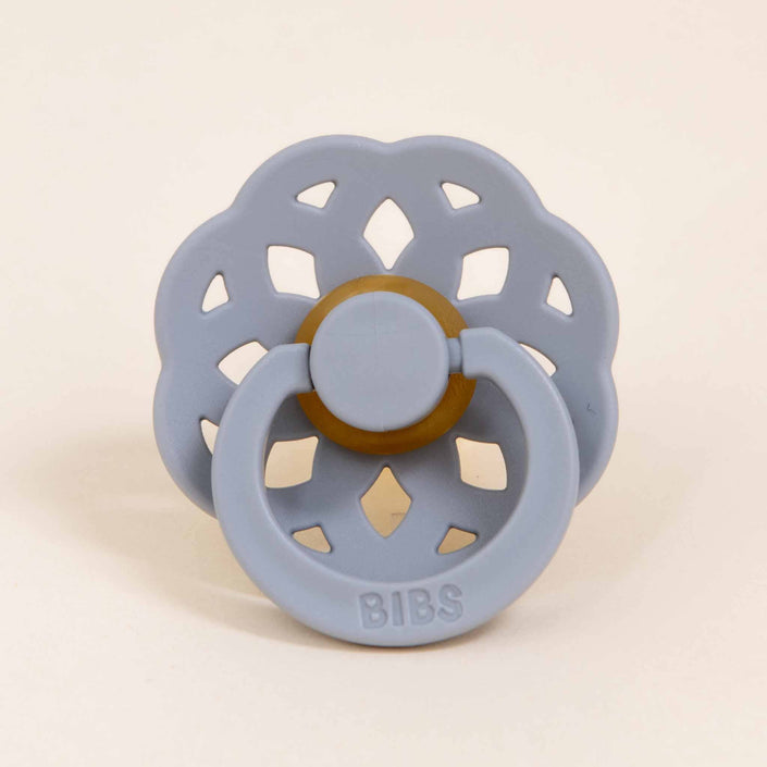 The Bibs Lace Pacifier in the color "Cloud" is positioned upright, showcasing its flower-shaped design. It includes a circular mouthpiece made from natural rubber and a decorative handle with cut-out petal shapes. Made in Denmark, the word "BIBS" is embossed on the bottom of the handle, encouraging your baby to naturally self-soothe.