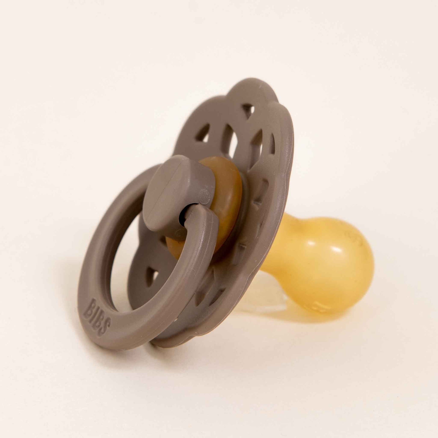 The Bibs Lace Pacifier in Dark Oak showcases a bohemian-inspired design, featuring a natural rubber construction with a round handle and perforated shield, all set against a neutral background. The soft yellow nipple is specially crafted to assist little ones in self-soothing.