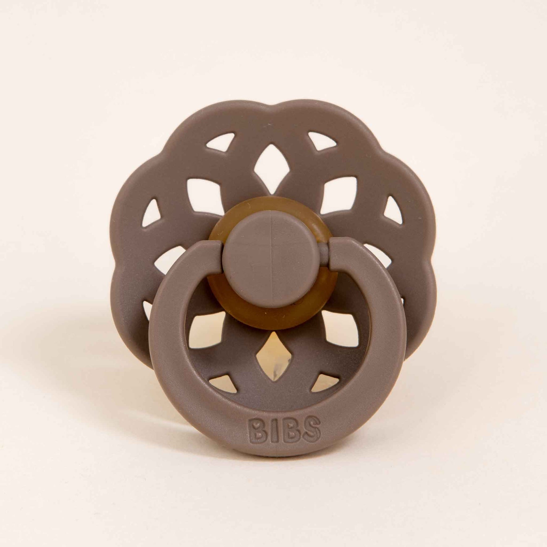 The Bibs Lace Pacifier in Dark Oak features a bohemian-inspired design with a flower-shaped shield and round handle made from natural rubber. The handle is embossed with the word "BIBS," set against a plain, light-colored background, making it an ideal choice for those looking to provide their baby with a gentle self-soothing experience.