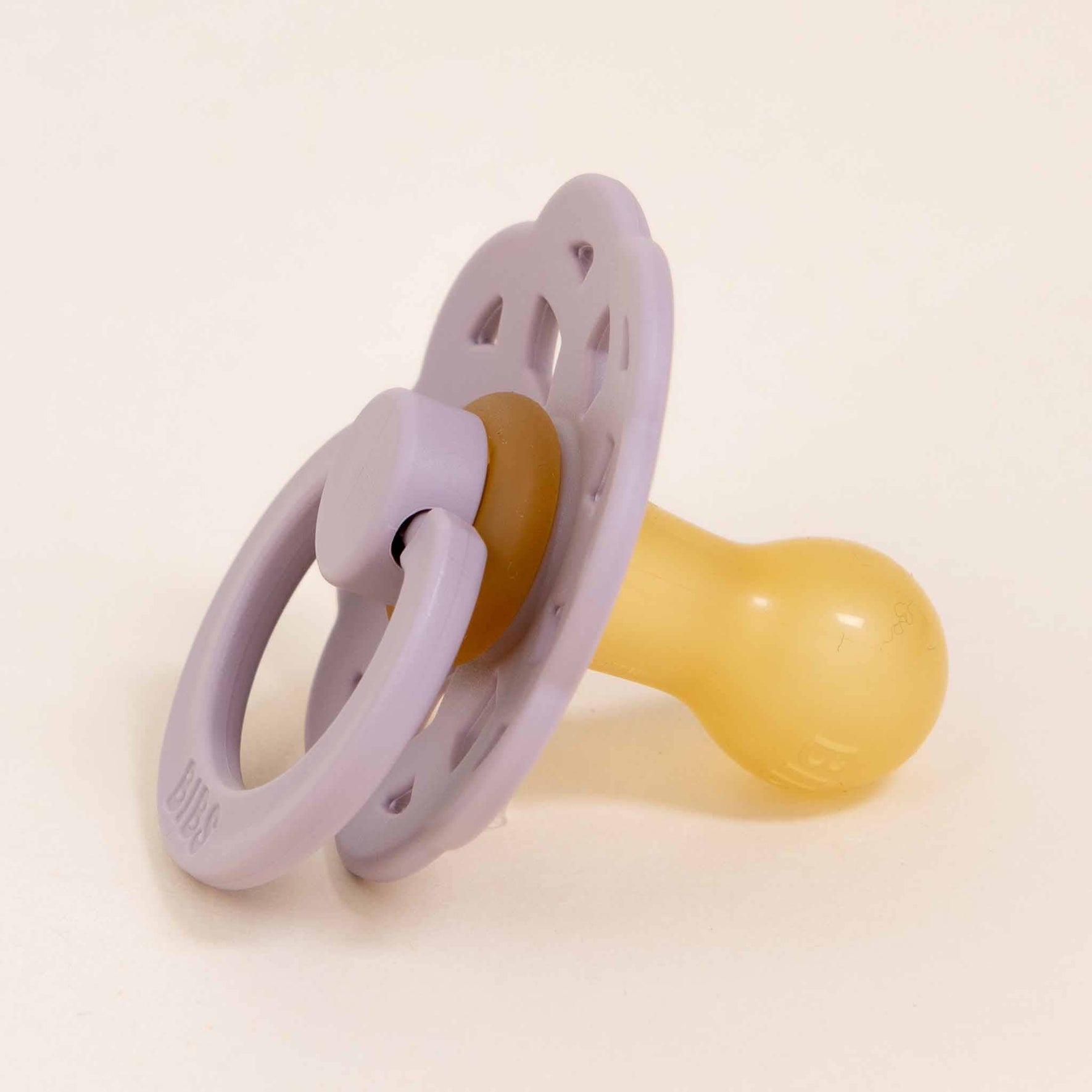 A Bibs Lace Pacifier from the Dusky Lilac 2 Pack, adorned with a bohemian-inspired lavender shield and ring and featuring a yellow silicone nipple, is placed on a light surface. Expertly crafted to help babies self-soothe in style.