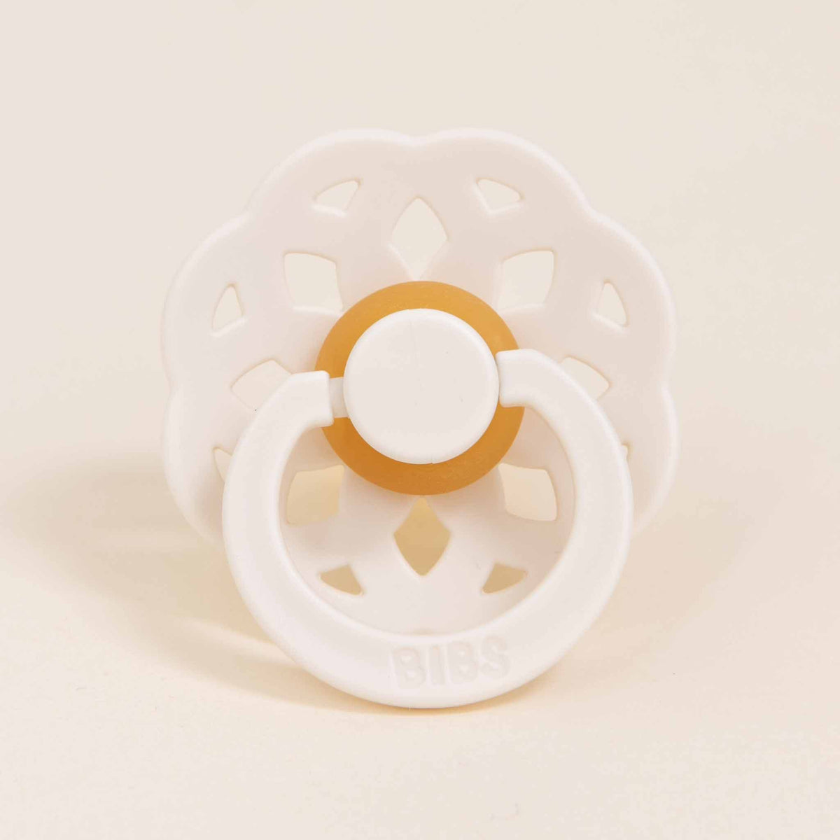 A Bibs Lace Pacifier in ivory, featuring a decorative, bohemian-inspired flower-shaped shield and a yellow ring, sits upright against a plain, light background. Made from natural rubber, the word "BIBS" is embossed on the pacifier, making it perfect for babies learning to self-soothe.