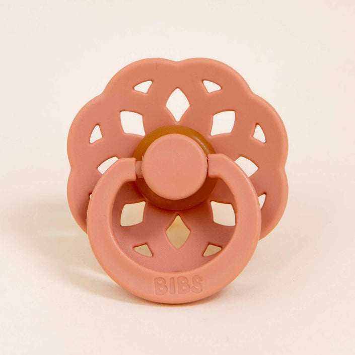 Displayed against a plain background is a peach, flower-shaped pacifier crafted from natural rubber. The bohemian-inspired design features cut-out petal sections and a circular handle embossed with "BIBS," showcasing its Danish origins. This is part of the Bibs Lace Pacifier 2 Pack.