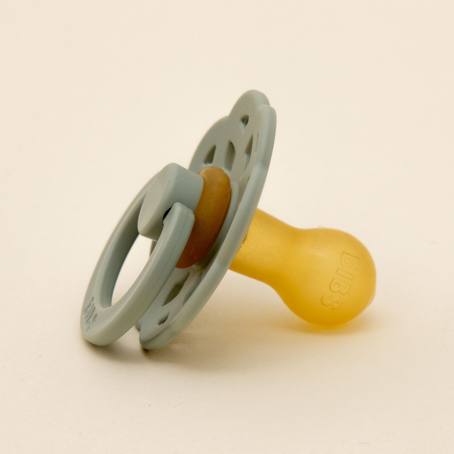 A Bibs Lace pacifier from the Sage set, featuring a light gray handle and shield with decorative holes, is showcased on a plain light background. Crafted in Denmark from natural rubber, the pacifier is positioned at an angle to highlight the brand name on its yellow surface.