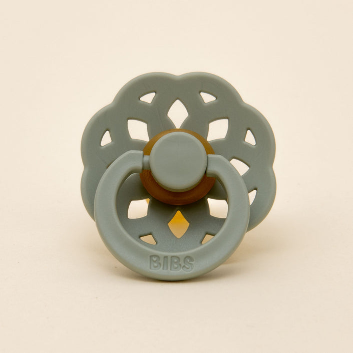 A pacifier from the Bibs Lace Pacifier 2 Pack in Sage, featuring a decorative bohemian-inspired floral shield design, stands upright against a plain light background. This natural rubber pacifier, made in Denmark, has the brand name "BIBS" embossed on its handle.