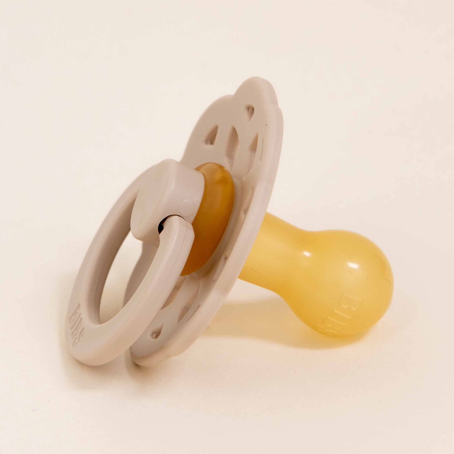Resting on a white surface is the Bibs Lace Pacifier 2 Pack in Vanilla, featuring a bohemian design on its beige and yellow pacifier with a ring handle. This natural rubber baby pacifier includes a round nipple and a circular shield with patterned holes, making it perfect for helping your little one self-soothe.