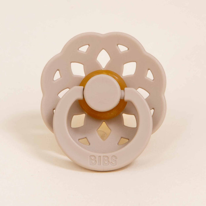 The Bibs Lace Pacifier 2 Pack in Vanilla features a unique beige pacifier with a floral-shaped base and a circular handle, embodying a bohemian design. The brand name "BIBS" is embossed on the handle, signifying its superior quality as a natural rubber baby pacifier. Ideal for little ones learning to self-soothe, it is displayed upright against a plain background.