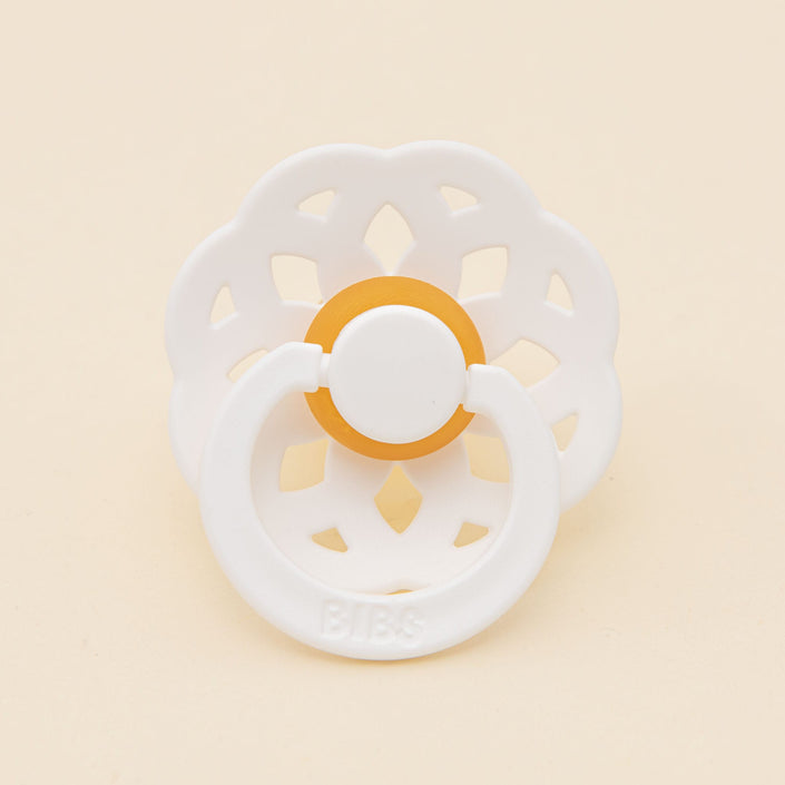 The Bibs Lace Pacifier from the 2-pack in White showcases a bohemian-inspired design with a floral-patterned shield and an orange accent, set against a plain, light background.