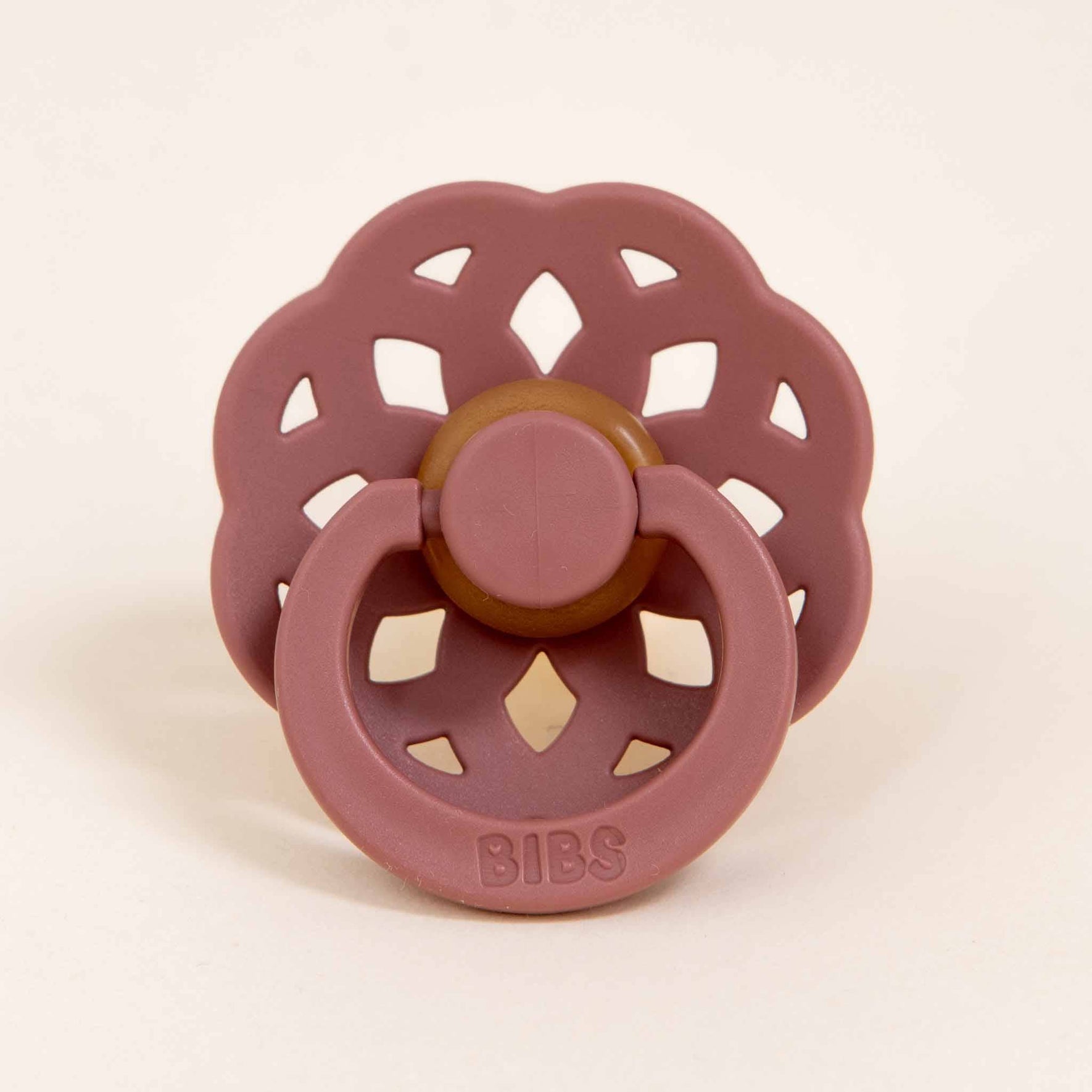 A pink Bibs Lace Pacifier from the Woodchuck 2 Pack, featuring a bohemian-inspired flower-shaped shield with a brown center, is placed on a light background. Made from natural rubber, the BIBS brand name is embossed on the handle, making it ideal for helping little ones self-soothe.